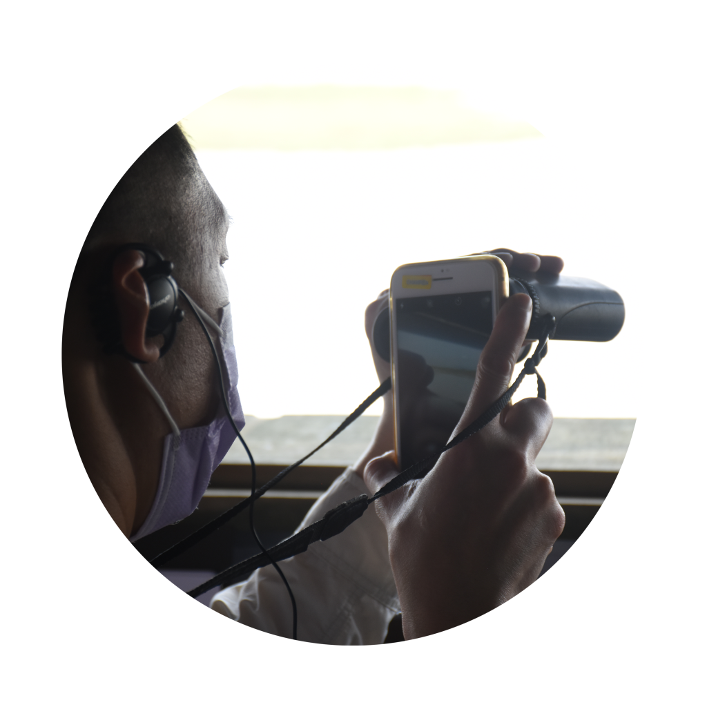 圓形圖片，在觀鳥屋窗邊的一位參加者一手提着望遠鏡、一手開着電話中的相機，嘗試以相機的鏡頭對焦望遠鏡中的影像。