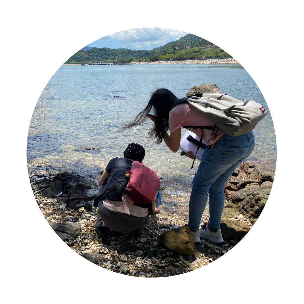圓形圖片，在海邊，有兩個背向鏡頭的團導賞參加者，其中視障參加者蹲下觸摸膝下的貝殼和石頭，她的同行者則在旁俯下身幫忙拍照。